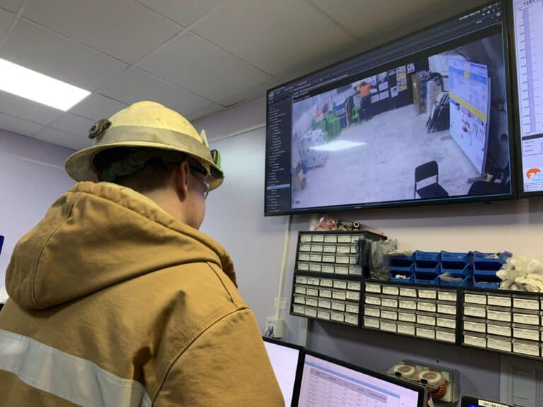 Mine worker in hard hat and high-visibility jacket monitoring underground refuge chamber via GuardIAN Intellegence system on large wall-mounted screen in control room